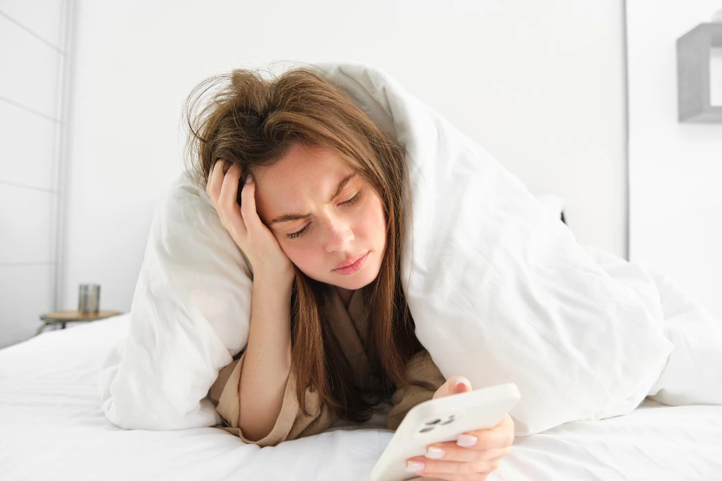 Depressed woman in bed lying under blanket and looking at phone with sad complicated face